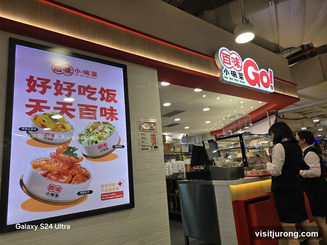 The storefront of a food establishment with a large illuminated advertisement on the left displaying three bowls of food and Chinese text. The main signage above the counter reads "Bai Wei Mini Bowl Go!" in English and Chinese. Two employees in uniform are visible behind the counter.