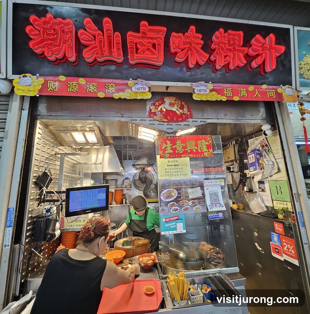 A food stall with a large red neon sign in Chinese characters above the entrance. Below the sign is a banner with smaller Chinese characters and NETS logos. The stall is open, showing a kitchen area with cooking equipment and staff preparing food. There are menus displayed inside the stall.