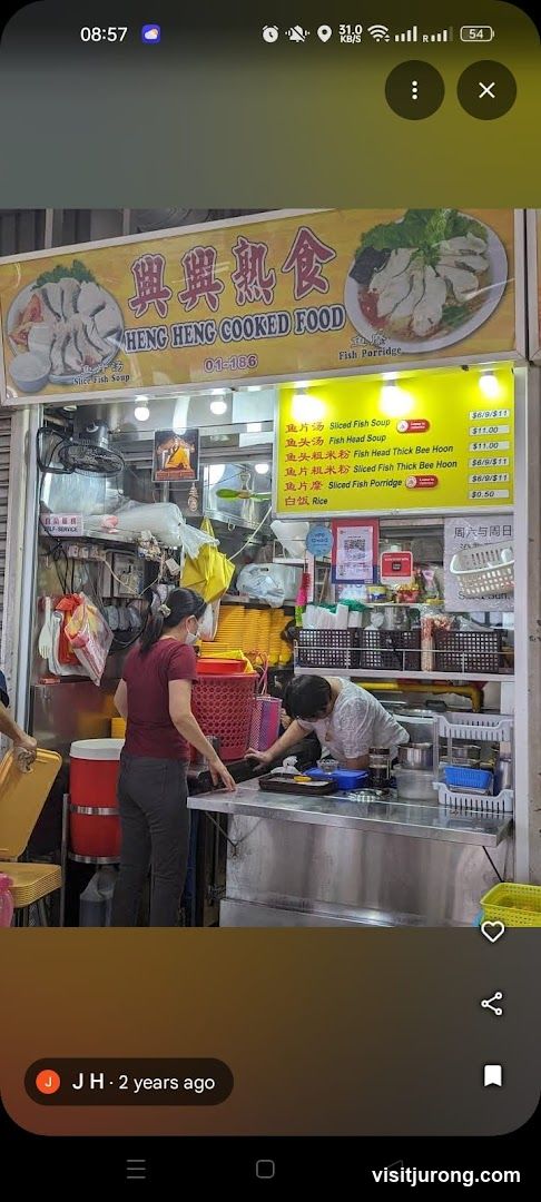 A food stall with a yellow awning displaying the name "HENG HENG COOKED FOOD" and images of fish soup and fish porridge. A menu is displayed below the awning. Two staff members are visible behind the counter, preparing food.