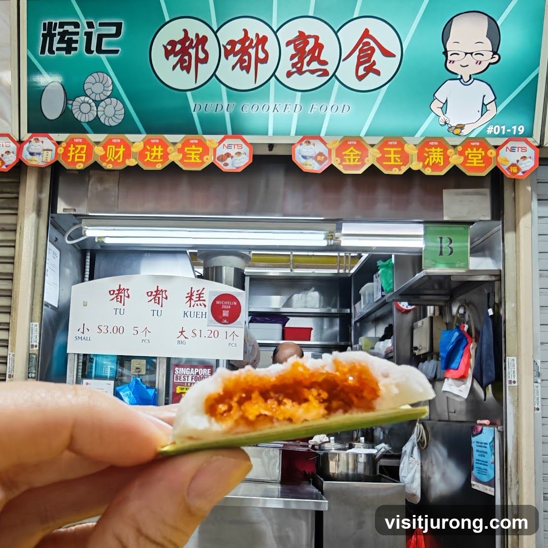 A close-up of a hand holding a Dudu Kueh in front of a food stall. The stall has a sign with Chinese characters and "DUDU COOKED FOOD" written below. There is also a price list for small and big Dudu Kueh. The stall appears to be a Michelin Bib Gourmand recipient.