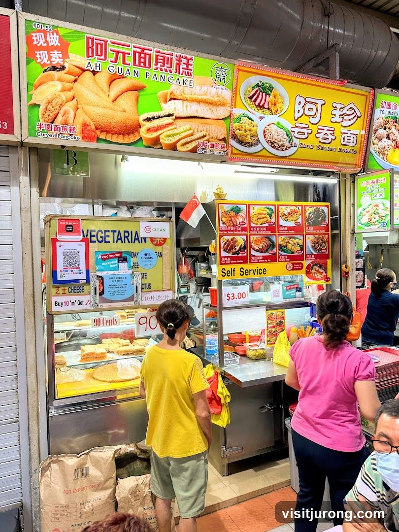 A food stall with two prominent signs. The left sign advertises "AH GUAN PANCAKE" with images of curved and sliced pancakes. The right sign advertises "Ah Zhen Wanton Noodle" with images of noodle dishes. Below the signs, the stall displays various food items in a refrigerated case, with a "VEGETARIAN" sign and pricing for cheese items. A "Self Service" counter with a $3.00 price is also visible. Two customers are at the counter.