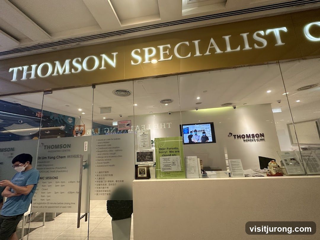 The exterior of a Thomson Specialist Clinic, featuring large glass doors and windows. Above the entrance, the words "THOMSON SPECIALIST C" are illuminated. Inside, a reception area is visible with signage for Thomson Paediatric Centre and Thomson Women's Clinic. A person wearing a mask is standing near the entrance.