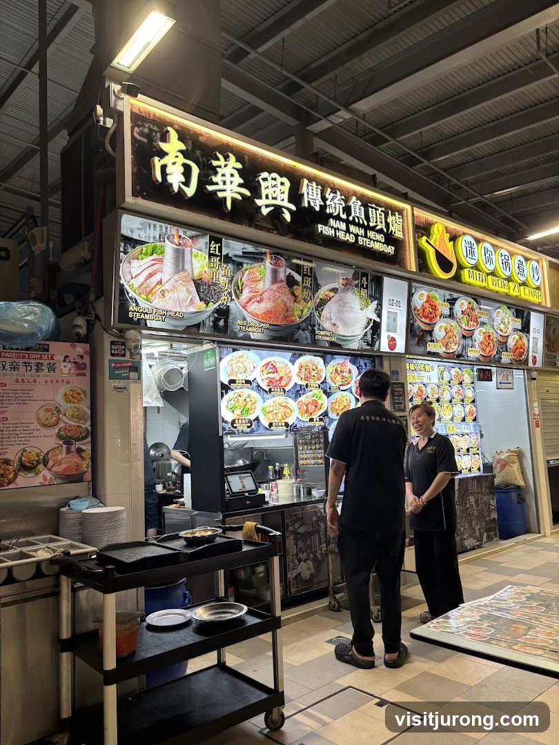 A brightly lit food stall with a large sign displaying the business name and images of various steamboat dishes. The sign features Chinese characters and English text. Below the main sign are illuminated panels showcasing different steamboat options like "Angoli Fish Head Steamboat" and "Red Grouper Steamboat." To the right, there's a section for "Mini Wok Zi Char" with pictures of individual meals. Two staff members are visible in front of the stall, and a trolley with plates and condiments is positioned to the left.