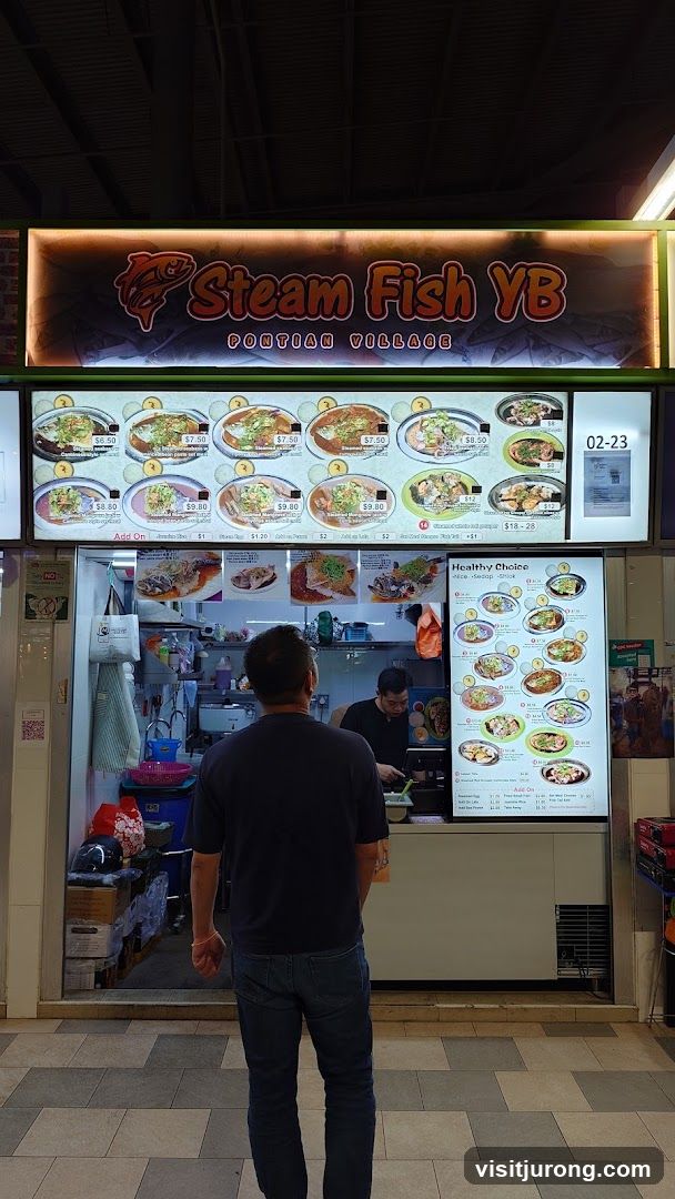 A food stall with a large illuminated sign displaying the business name and various dishes. Below the sign is a menu board with pictures and prices of steamed fish dishes. A customer is standing in front of the counter, and a staff member is visible behind it.