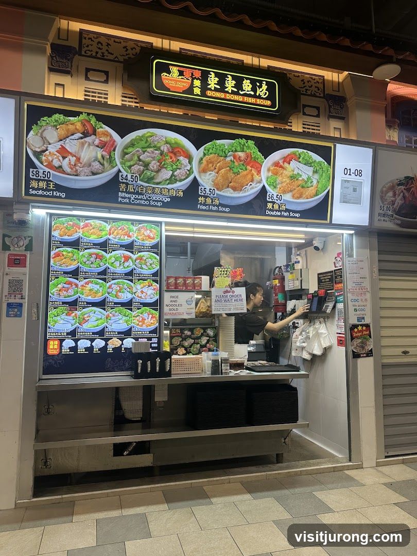 A food stall with a prominent sign displaying "DONG DONG FISH SOUP" and images of various fish soup dishes. The stall features a menu board with prices and descriptions of the dishes, including Seafood King, Bittergourd/Cabbage Pork Combo Soup, Fried Fish Soup, and Double Fish Soup. A staff member is visible behind the counter, attending to a customer or order. The stall is located in an outdoor or semi-outdoor food court setting.