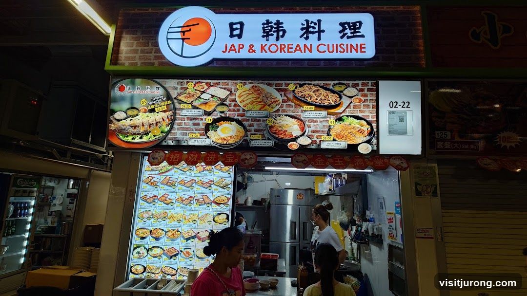 A storefront for a Japanese and Korean cuisine restaurant. The facade features a large illuminated sign with the restaurant's name and logo, along with numerous food images and menu items displayed. The interior of the kitchen is visible, with staff members preparing food.