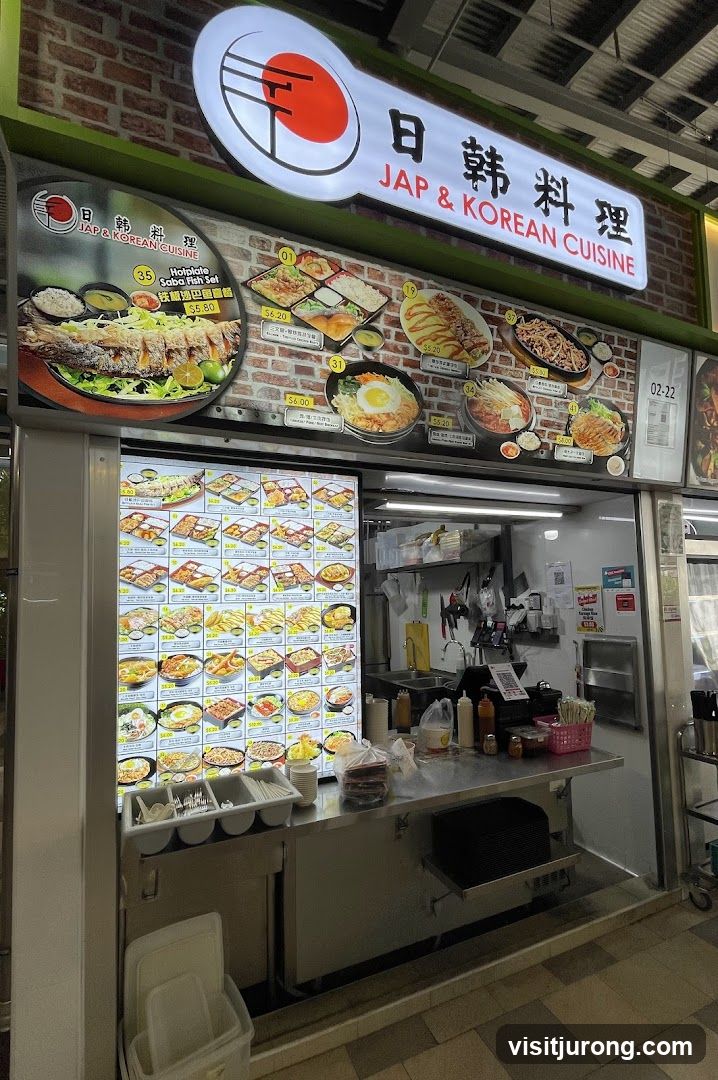 A food stall with a large illuminated menu displaying various Japanese and Korean dishes. The stall has a prominent sign at the top that reads "JAP & KOREAN CUISINE" in English and Chinese characters. Below the sign, a large display shows pictures and prices of food items. The counter area has condiments and utensils.