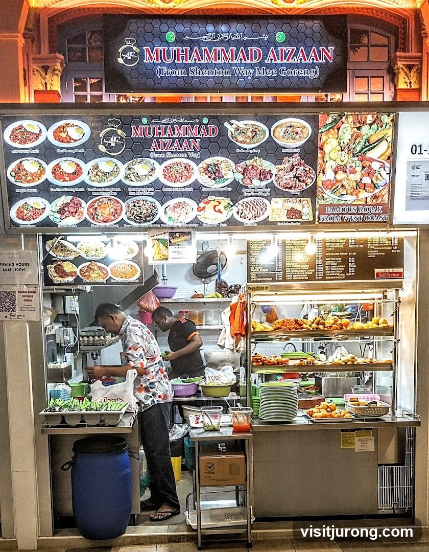 A food stall with a large menu board displaying various dishes and their images. Two men are working behind the counter. The stall has a sign with the name "MUHAMMAD AIZAAN" and "(From Shenton Way Mee Goreng)". Below the name are pictures of different food items. To the right, there is a section for "FAMOUS ROJAK FROM WEST COAST". A menu board with prices is visible on the right side.