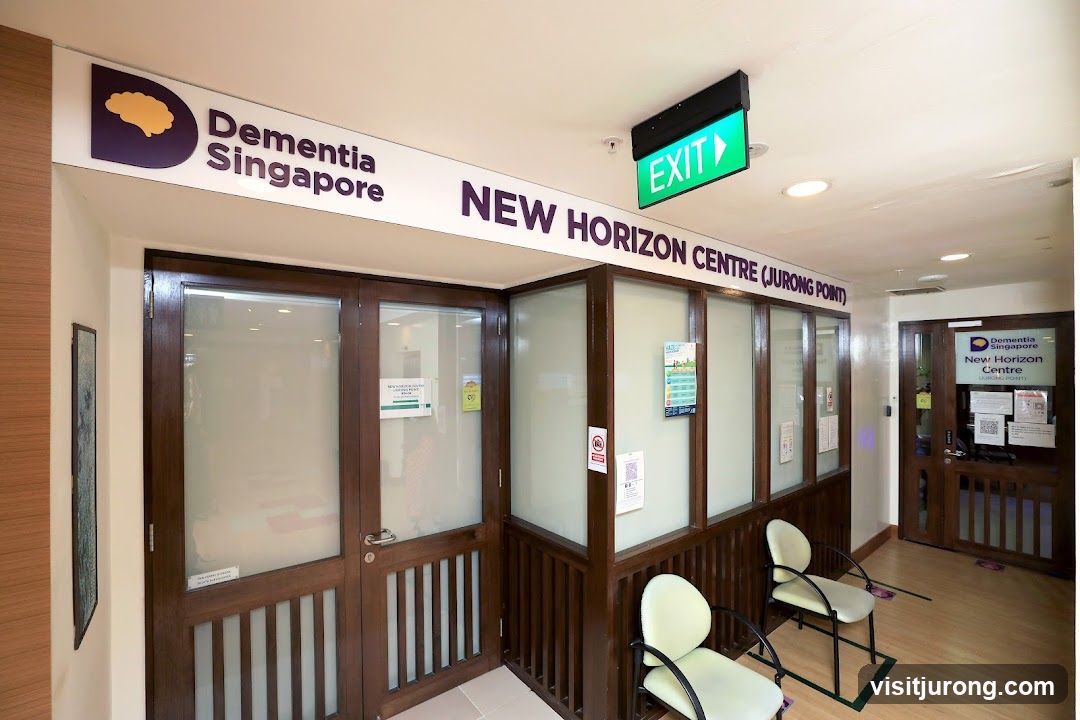 The exterior of the New Horizon Centre (Jurong Point) is visible, with signage for Dementia Singapore above the entrance. The entrance features double wooden doors with frosted glass panels. Inside, chairs are arranged in a waiting area. An exit sign is illuminated above the entrance.