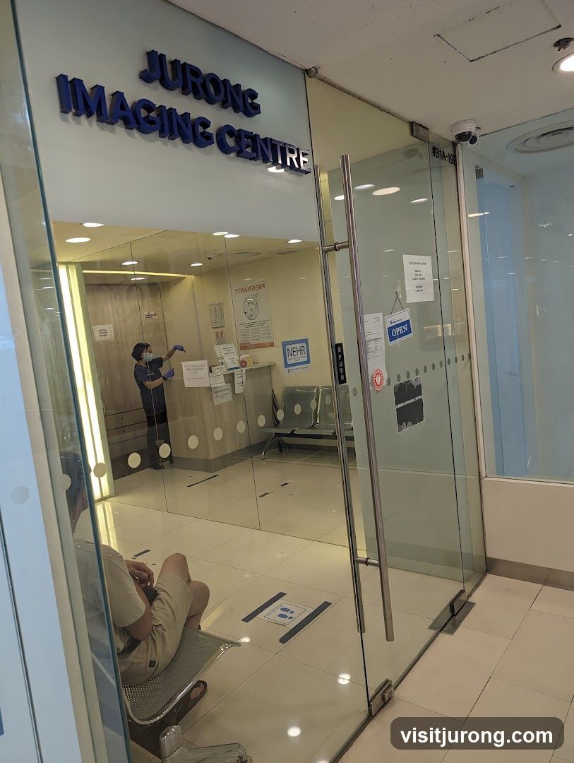 The exterior of Jurong Imaging Centre, a medical facility. The entrance features glass doors and windows with the facility's name prominently displayed above the entrance. Inside, a waiting area with chairs is visible, along with a staff member attending to a counter. Social distancing markers are on the floor.