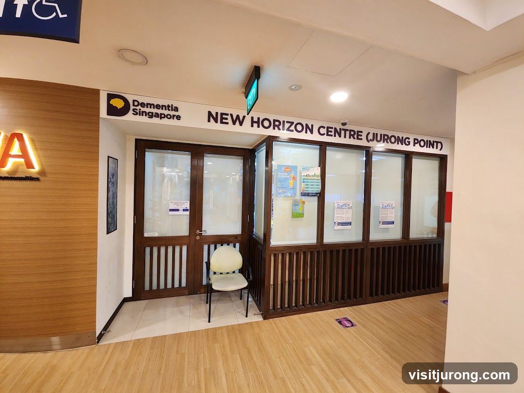 The exterior of the New Horizon Centre (Jurong Point) is visible, with a sign above the entrance that reads "Dementia Singapore NEW HORIZON CENTRE (JURONG POINT)". The entrance has double wooden doors and large glass windows. A single chair is placed outside the entrance. There are posters displayed on the windows.