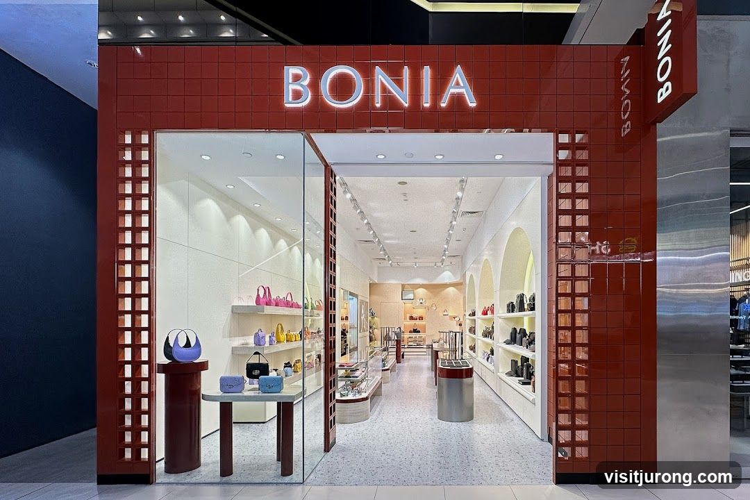 The exterior of a Bonia store, featuring a red tiled facade with the store name "BONIA" in illuminated letters above the entrance. The store has a large glass window display showcasing various handbags and accessories. Inside, the store is brightly lit with shelves displaying more merchandise, including handbags and shoes. The interior has a modern and clean aesthetic with arched displays and a speckled floor.