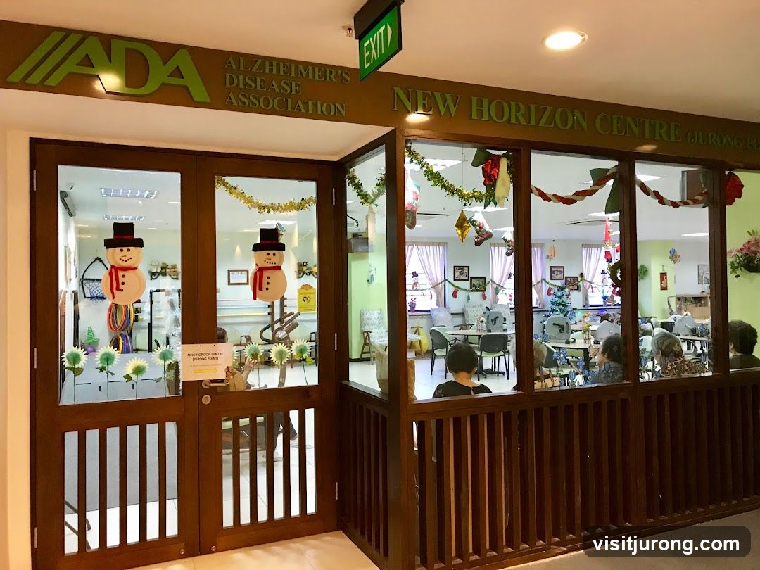The exterior of the New Horizon Centre, part of the Alzheimer's Disease Association. The entrance features double wooden doors with glass panels decorated with snowman cutouts and floral motifs. Above the entrance, signage reads "ADA ALZHEIMER'S DISEASE ASSOCIATION" and "NEW HORIZON CENTRE (JURONG POINT)". The interior is visible through the glass, showing a room decorated for the holidays with a Christmas tree and festive garlands. Several people are seated at tables inside.