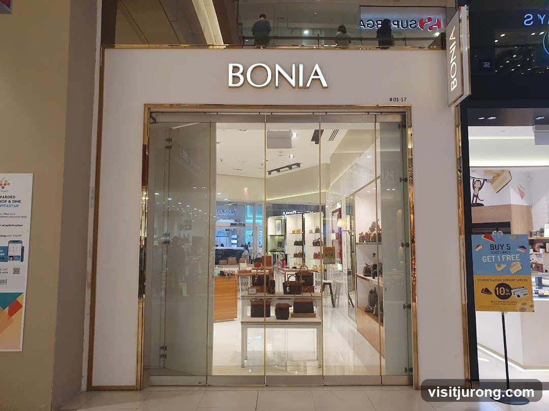 The exterior of a BONIA store with large glass doors and windows. The store name "BONIA" is prominently displayed in gold lettering above the entrance. Inside, handbags and accessories are displayed on shelves and stands. A promotional sign for "BUY 5 SLICE CAKES GET 1 FREE" is visible to the right of the entrance.