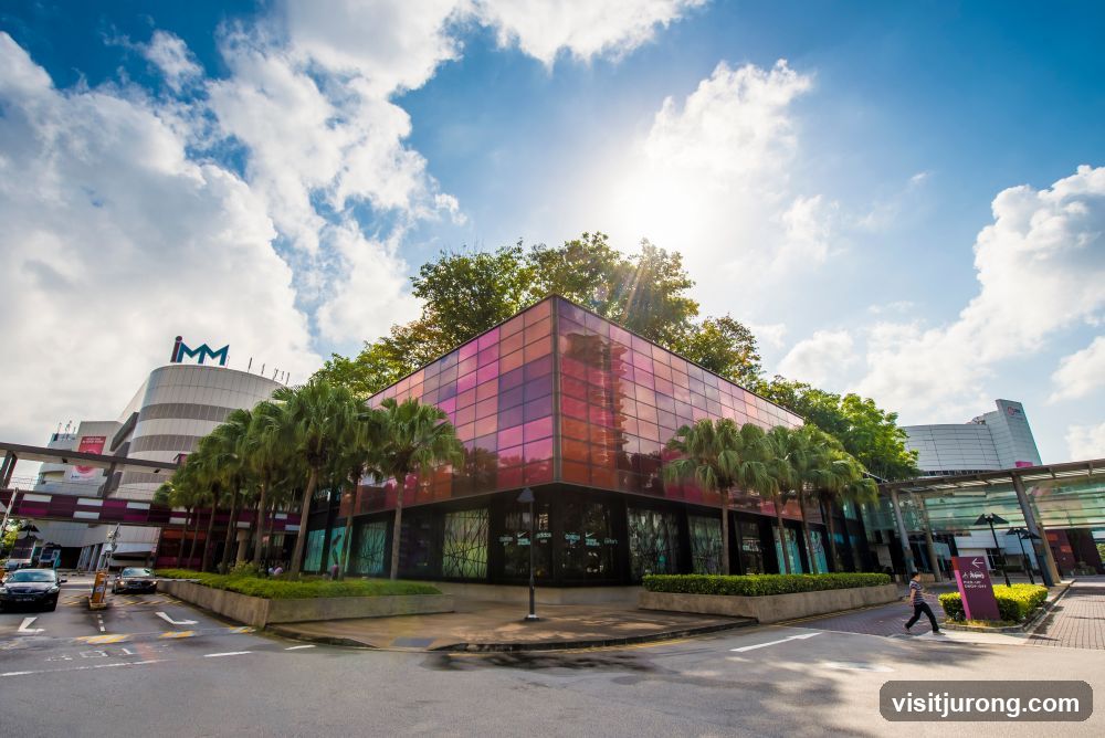 A modern building with a colorful glass facade, surrounded by palm trees and a bright blue sky with fluffy clouds. The IMM logo is visible on a taller adjacent building.