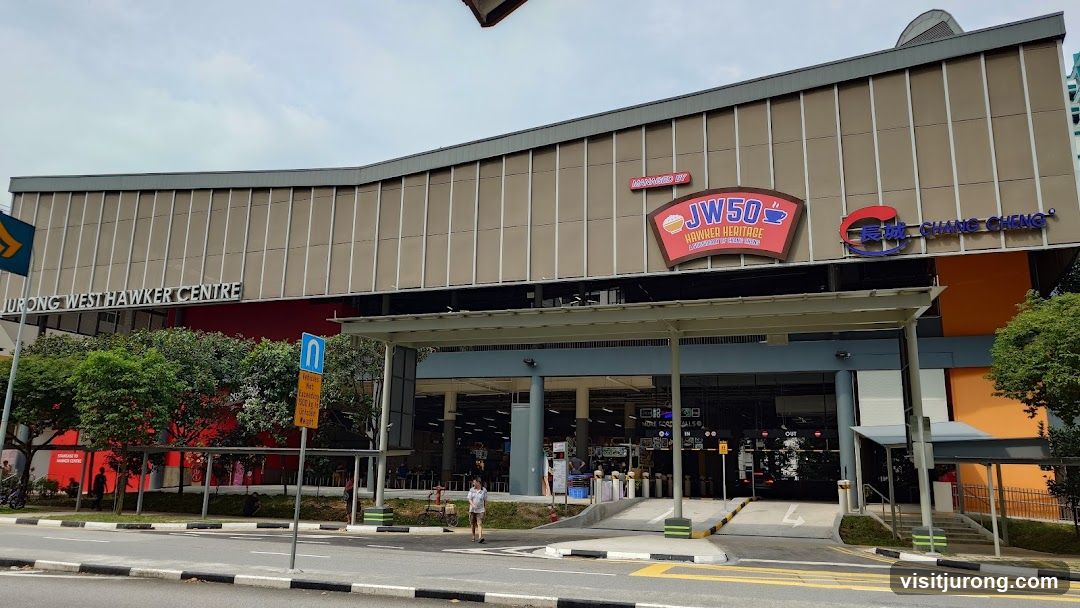The exterior of a hawker centre with multiple signs visible. The main sign reads "JURONG WEST HAWKER CENTRE". Above the entrance, there are signs for "JW50 HAWKER HERITAGE" and "CHANG CHENG". The building has a modern facade with large windows and covered walkways.