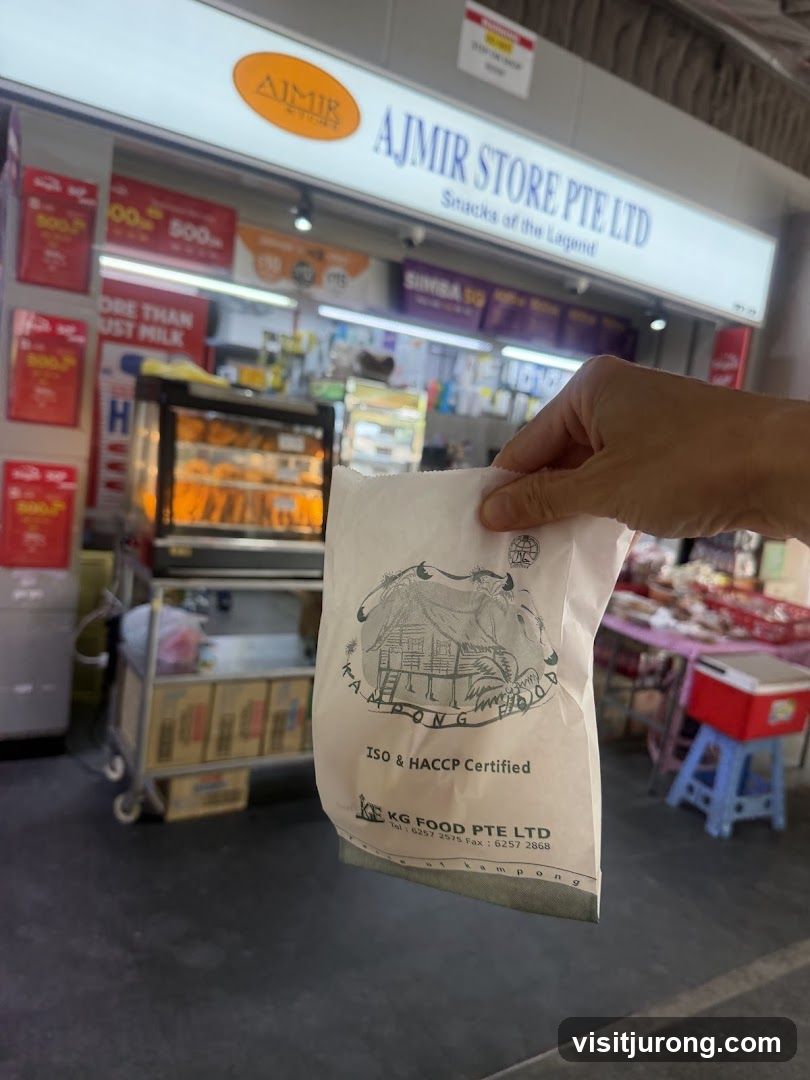 The exterior of Ajmir Store Pte Ltd, a shop selling snacks. The store has a large illuminated sign with the store name and "Snacks of the Legend". There are also promotional posters and a display fridge with food items. A person is holding a paper bag with a logo and text that reads "KG FOOD PTE LTD".