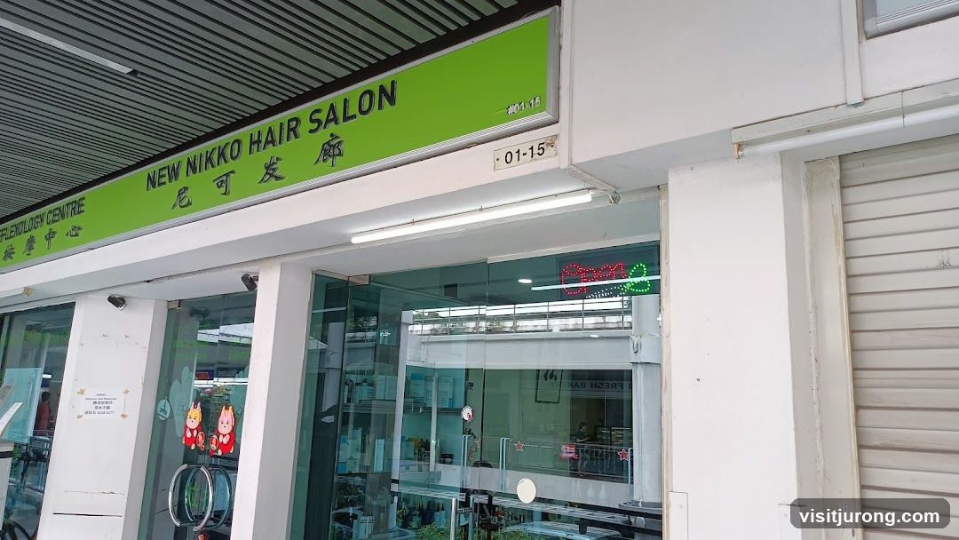 The exterior of a hair salon with a green sign that reads "NEW NIKKO HAIR SALON" in English and Chinese characters. Below the sign, the unit number "01-15" is visible. The storefront features large glass windows and doors, with an "Open" sign illuminated in neon lights inside. There are also some decorative figures on the glass doors. To the left of the salon, a sign for "REFLEXOLOGY CENTRE" is partially visible.