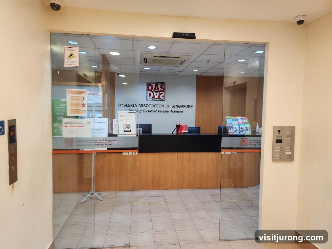 The entrance to the Dyslexia Association of Singapore learning centre. The main signage with the logo and name is visible on the wall behind the reception desk. The entrance is a glass door with various notices and QR codes displayed on it.