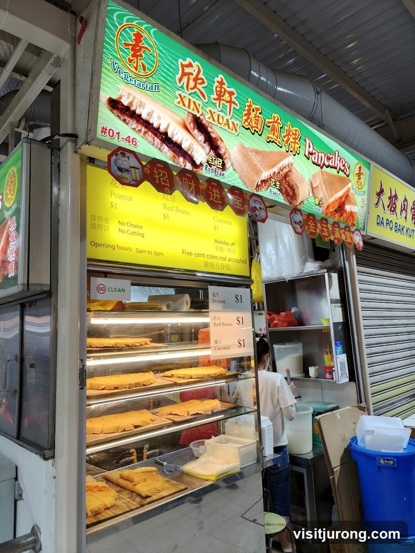 A street food stall selling vegetarian pancakes. The stall has a large illuminated sign with images of pancakes and the name "XIN XUAN" and "Vegetarian Pancakes". Below the sign is a menu board listing prices for different fillings. The stall displays several rows of freshly made pancakes in a glass display case. A person is visible behind the counter preparing food.