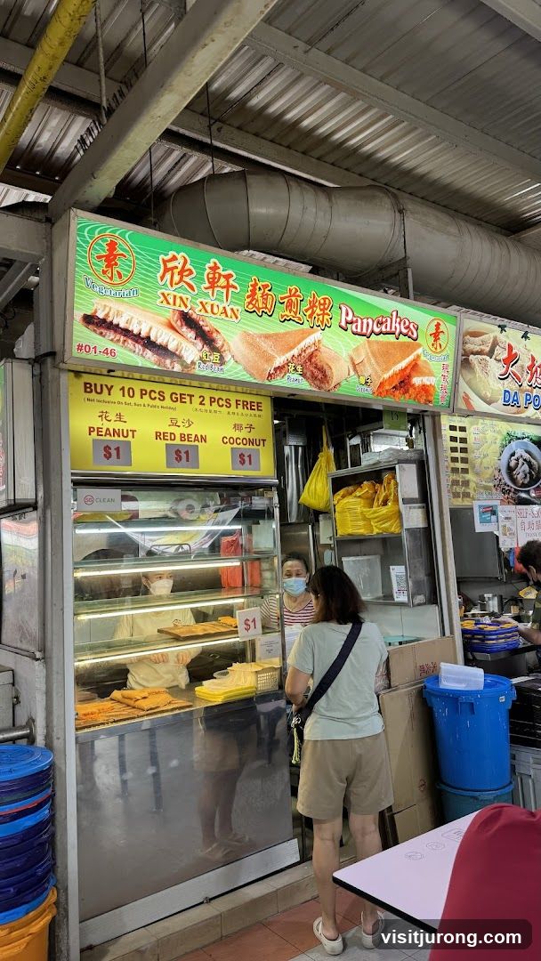A food stall with a prominent sign displaying "XIN XUAN Pancakes" and images of their products. Below the main sign is a yellow board advertising a promotion: "BUY 10 PCS GET 2 PCS FREE". It also lists the prices for Peanut, Red Bean, and Coconut pancakes at $1 each. A display case shows the pancakes, and two staff members are visible behind the counter.