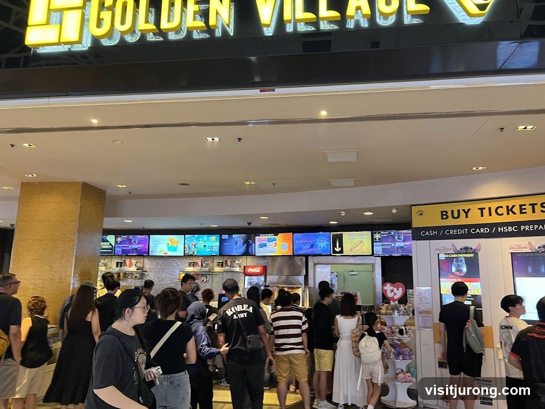 A view of the Golden Village cinema entrance with a line of people waiting. Above the entrance, the Golden Village logo is visible. To the right, a "BUY TICKETS" sign is displayed with payment options. Below the sign, ticket machines are present. To the left, digital screens show movie posters and F&B promotions.