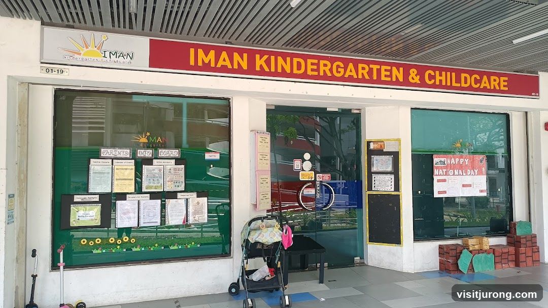 The storefront of IMAN Kindergarten & Childcare features a large red sign with yellow lettering above the entrance. The windows display various notices and decorations, including a "Parents Info Board" and a "Happy National Day" poster. A stroller is parked outside the entrance.