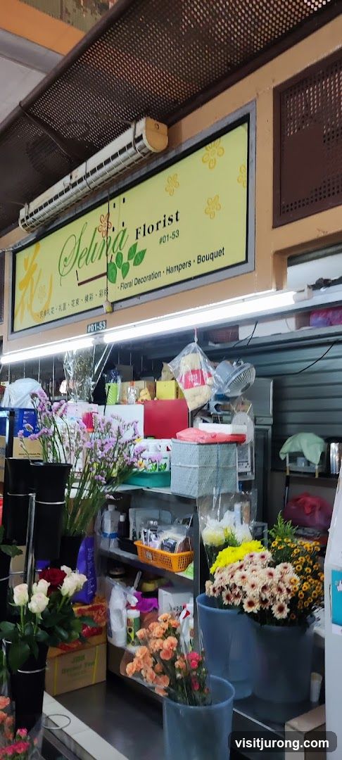 A florist shop with a sign that reads "Selina Florist #01-53 Floral Decoration, Hampers, Bouquet". The shop displays various flowers, including roses, chrysanthemums, and other assorted blooms, in buckets and vases. Shelves behind the flowers hold boxes, baskets, and other floral arrangement supplies.