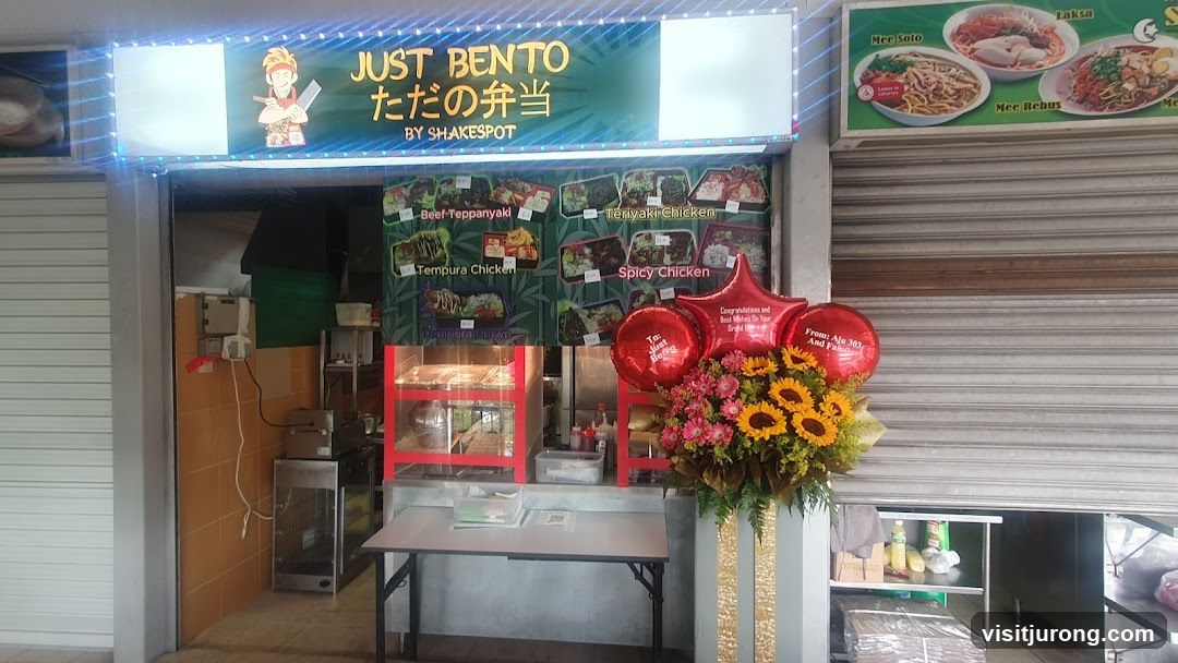 The exterior of a bento shop with a sign that reads "JUST BENTO ただの弁当 BY SHAKESPOT". Below the sign is a menu board displaying various bento options like Beef Teppanyaki, Teriyaki Chicken, Tempura Chicken, and Spicy Chicken. There are also images of Laksa, Mee Soto, and Mee Rebus on a separate sign. Red balloons and a floral arrangement are placed in front of the shop.