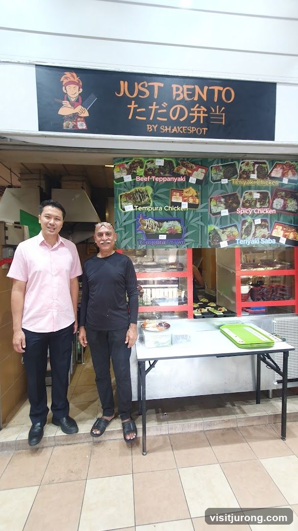 A storefront with a sign that reads "JUST BENTO ただの弁当 BY SHAKESPOT". Below the sign is a menu display featuring various bento box options. Two men are standing in front of the counter.