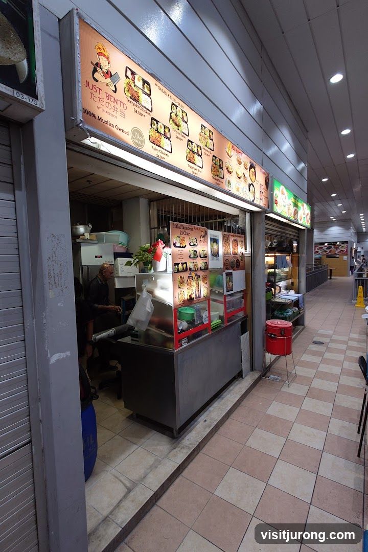A food stall with a large illuminated sign displaying bento box options. The stall is open, showing a counter with food displays and a person working inside. The surrounding area appears to be a food court or market with tiled flooring and other stalls visible in the background.