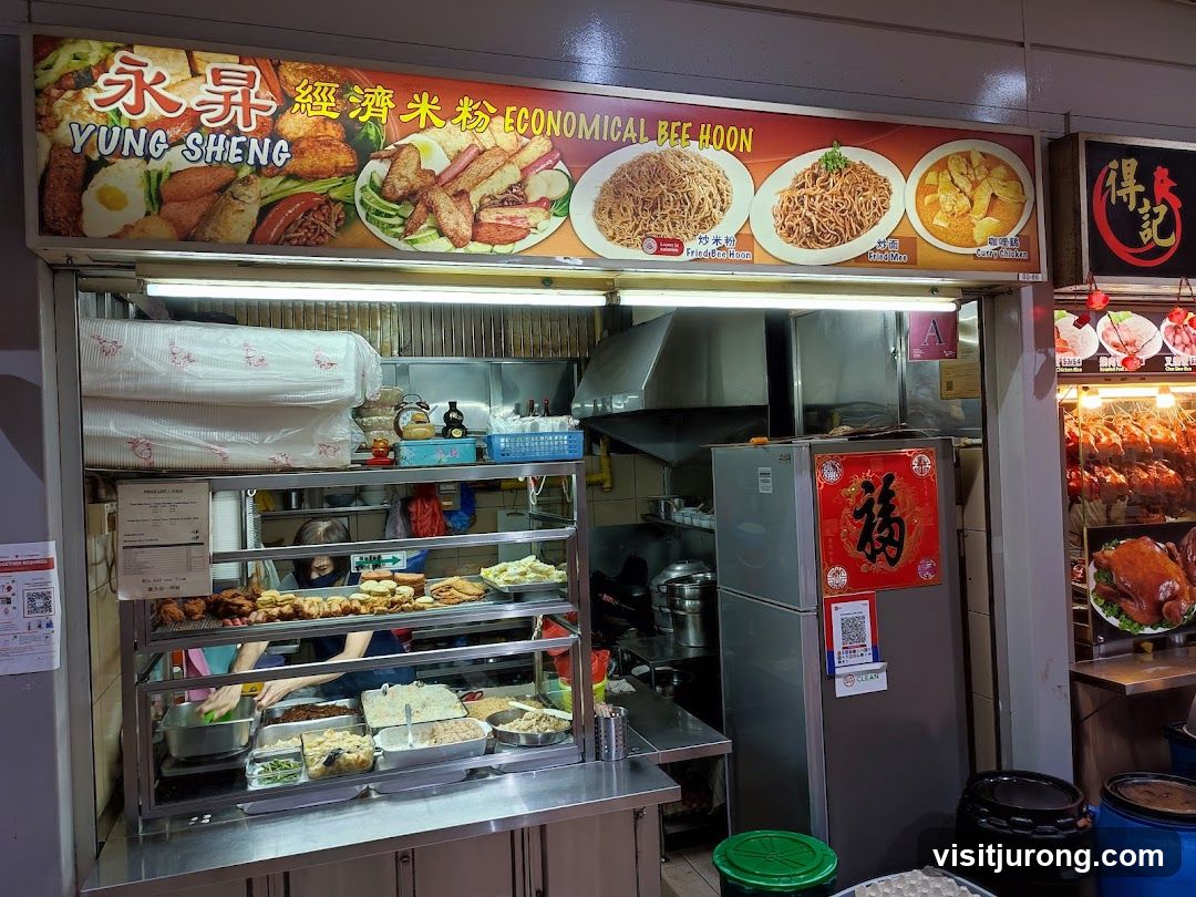 A food stall with a large sign displaying images of various dishes and the name "YUNG SHENG ECONOMICAL BEE HOON". Below the sign, food items are displayed in a refrigerated case, and a person is seen preparing food. To the right, roasted chickens are displayed.
