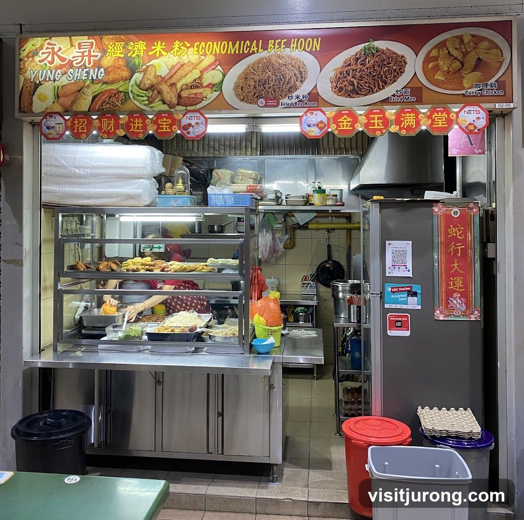 A food stall with a display of fried food items, noodles, and curry chicken. The stall has a sign with the name "YUNG SHENG ECONOMICAL BEE HOON" and images of the food offered. A person is seen preparing food behind the counter.