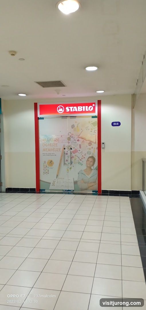 The exterior of a Stabilo store with a prominent red and white sign above the entrance. The glass doors and windows are decorated with graphics related to stationery and learning, including text that says "ORGANIZE COLORIZE MEMORIZE" and an image of a young woman. A unit number, "104-04", is visible on the wall to the right of the entrance.