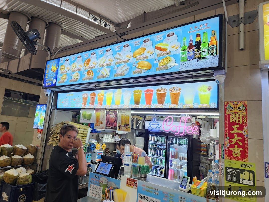 A brightly lit coffee stall with a large digital menu board displaying food and drinks. The stall has a neon "Coffee" sign and a refrigerator stocked with beverages. Coconuts are piled up outside the stall.