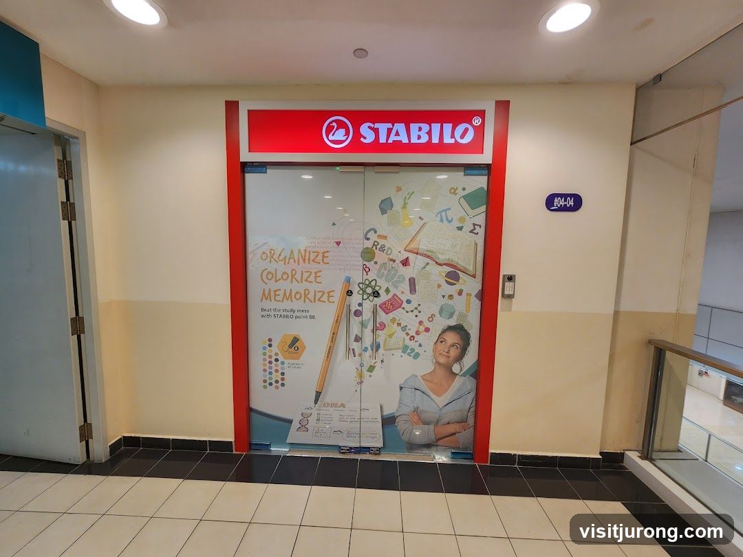 The storefront of a Stabilo store features a red and white illuminated sign with the Stabilo logo and name. The glass doors are decorated with graphics related to studying and art supplies, including the text "ORGANIZE COLORIZE MEMORIZE" and an image of a woman looking upwards. A number plate '#04-04' is visible on the wall next to the entrance.