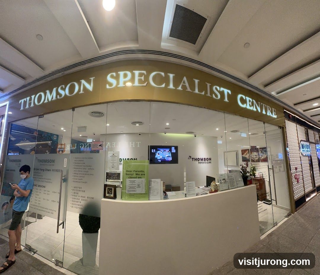 The exterior of a medical clinic with large glass windows and a prominent sign that reads "THOMSON SPECIALIST CENTRE". Inside, a reception desk is visible with staff and various informational posters. A person in a blue shirt and mask is standing near the entrance, looking at their phone.