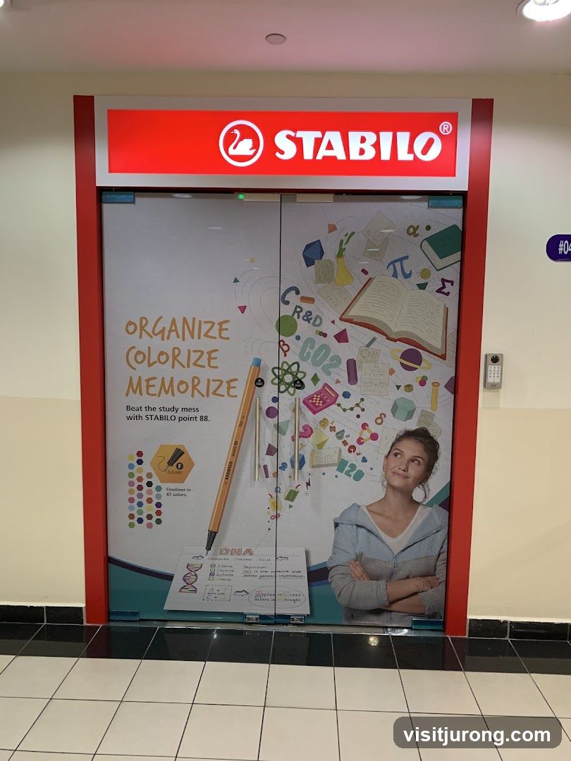 The image shows the storefront of a Stabilo store. The entrance is a double glass door with a large graphic wrap. The graphic features educational and scientific imagery, including DNA diagrams, chemical formulas, and mathematical symbols. A large Stabilo point 88 pen is prominently displayed. Text on the graphic reads "ORGANIZE COLORIZE MEMORIZE" and "Beat the study mess with STABILO point 88." A woman is depicted looking upwards, seemingly inspired. Above the doors, a red illuminated sign displays the Stabilo logo and name.