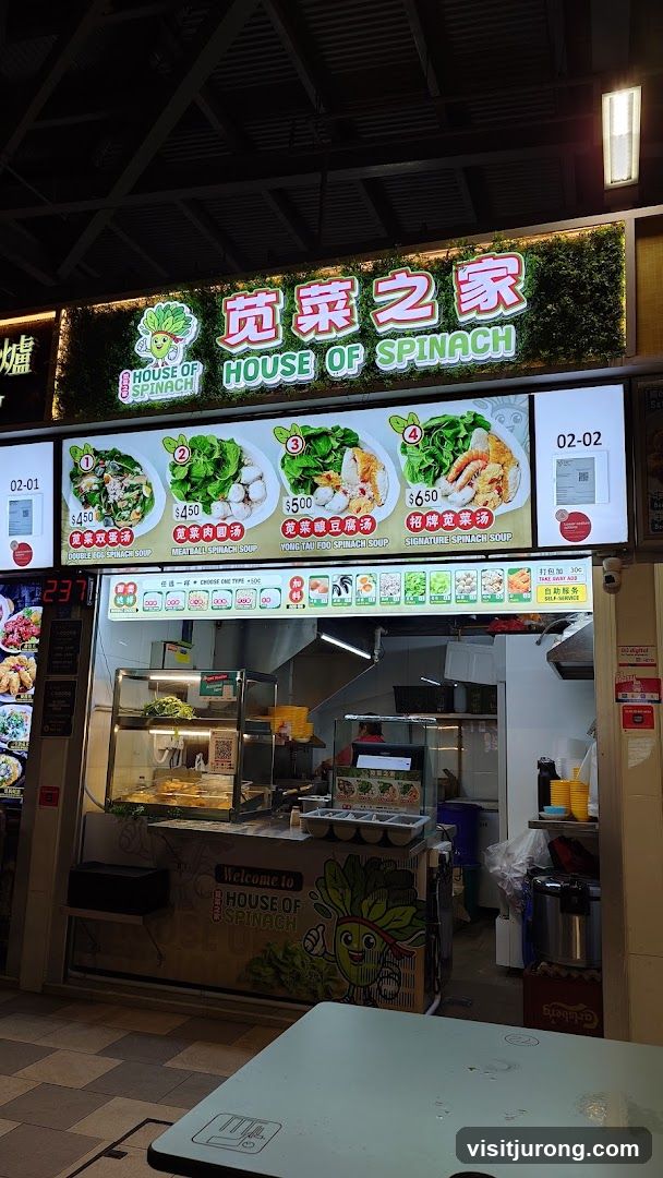 A food stall with a large illuminated sign that reads "HOUSE OF SPINACH" in English and Chinese characters. Below the main sign, there are pictures and prices for four different spinach soup dishes. The stall has a counter with food displayed and a menu board above.
