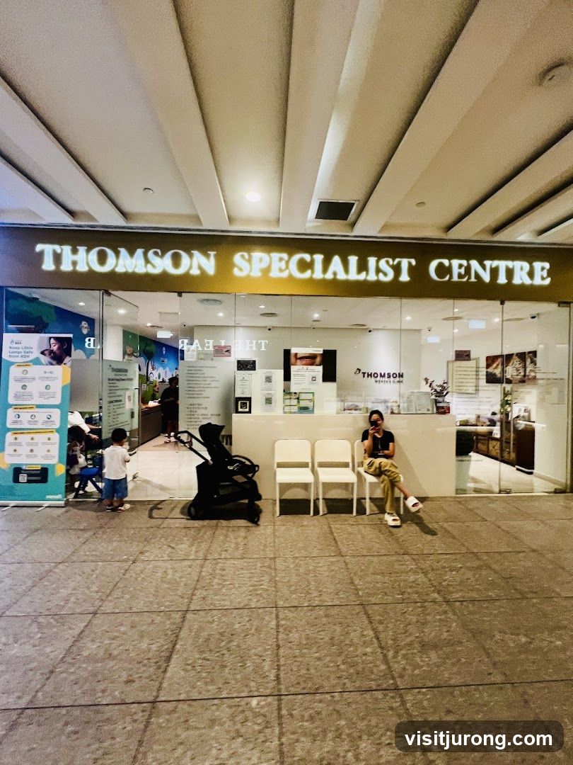The exterior of a medical clinic with a large sign reading "THOMSON SPECIALIST CENTRE" above the entrance. Inside, a reception desk is visible, along with several chairs and a stroller. A woman is seated in one of the chairs.