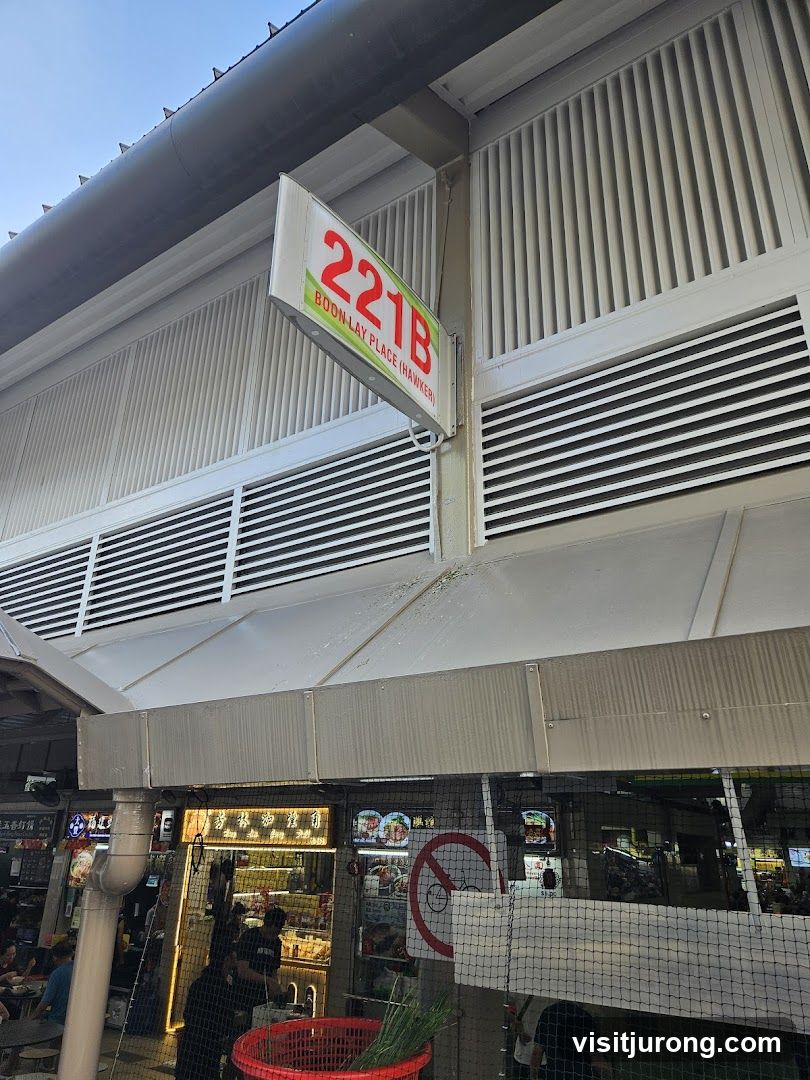 A sign for 221B Boon Lay Place (Hawker) is visible above a row of food stalls. The stalls have various signs and displays of food. A red basket with green onions is in the foreground.