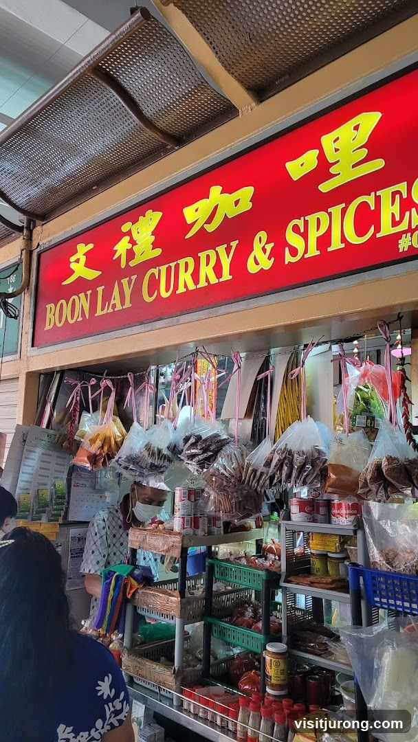 A red sign with yellow Chinese characters and the text "BOON LAY CURRY & SPICES" is displayed above a stall. Below the sign, various food items and spices are displayed in plastic bags and containers. A person wearing a mask is visible behind the counter.
