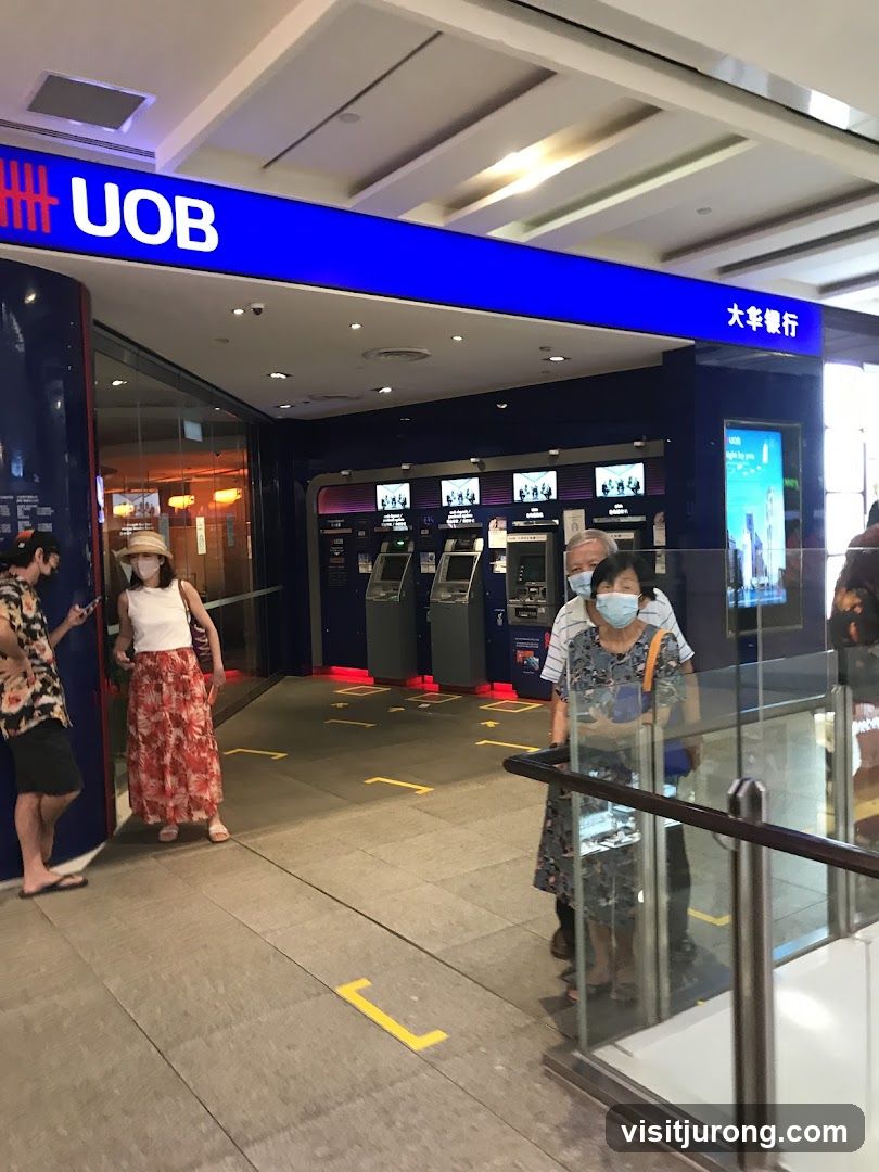 The exterior of a UOB bank branch with a prominent blue sign displaying the UOB logo and name. Several ATMs are visible inside, along with social distancing markers on the floor. People are seen queuing or standing near the entrance.