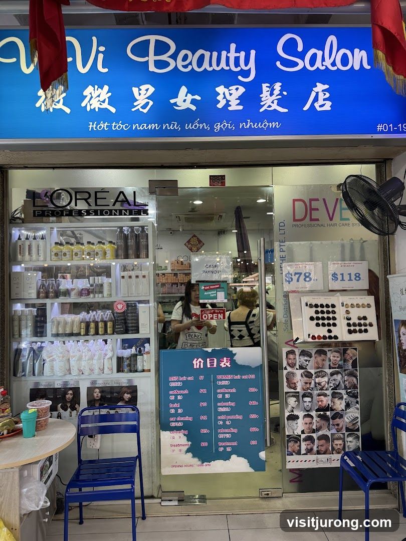 The exterior of a beauty salon with a large blue sign that reads "Vi Beauty Salon" in English and Chinese characters. Below the main sign, there is text in Vietnamese. The storefront has large glass windows displaying shelves of hair products and a price list. A woman is visible inside the salon.