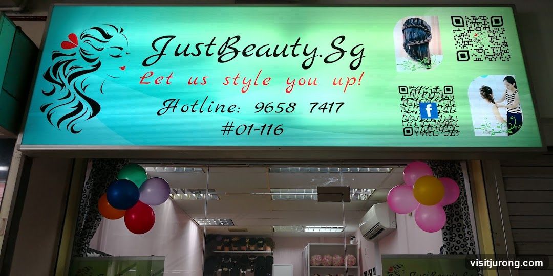 A brightly lit sign above a storefront displays the business name "JustBeauty.Sg" with the tagline "Let us style you up!". It also shows a hotline number and unit number. Images of hairstyles and QR codes are present on the sign. Balloons are arranged on either side of the entrance.