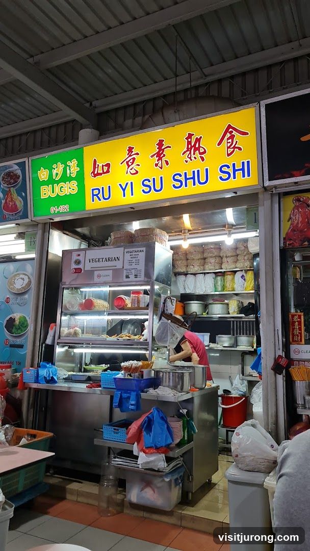 A vegetarian food stall with a bright yellow sign displaying Chinese characters and "RU YI SU SHU SHI" in English. Below the sign, the stall is visible with food displayed in a refrigerated case and shelves stocked with ingredients and supplies. A person in a pink shirt is working behind the counter.