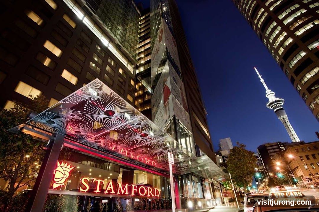 The exterior entrance of the Stamford Hotel at night. The hotel features a modern glass canopy with decorative patterns and neon red signage that reads "STAMFORD". The surrounding buildings are tall and illuminated, and the Sky Tower is visible in the distance. A taxi with "VIP CABS" signage is parked on the street.
